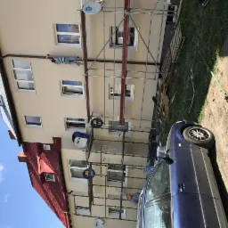 Praca na rusztowaniu przy elewacji budynku wielorodzinnego. Widoczni pracownicy. Budynek ocieplony, pomalowany na beżowo, z czerwonym dachem. Ujęcie z dołu.