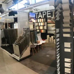 Ekspozycja w sklepie z tapetami i materiałami wykończeniowymi, prezentująca wzory tapet, listwy przypodłogowe, panele podłogowe i inne materiały dekoracyjne, z widocznym napisem 'TAPET'.
