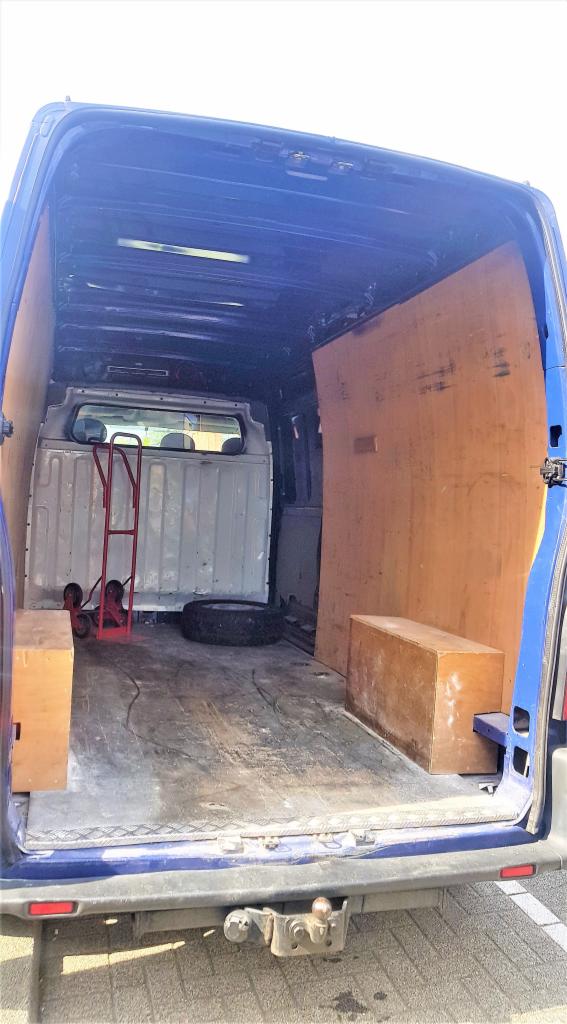 Wnętrze niebieskiego busa z drewnianą zabudową, widoczna opona, wózek transportowy i kartonowe pudła, hak holowniczy na zewnątrz pojazdu.