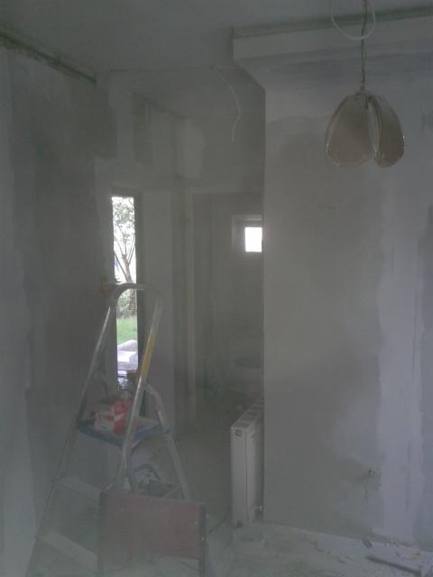 Pokój w trakcie remontu: ściany po szpachlowaniu, drabina malarska z farbą, wisząca lampa bez klosza, grzejnik centralnego ogrzewania.