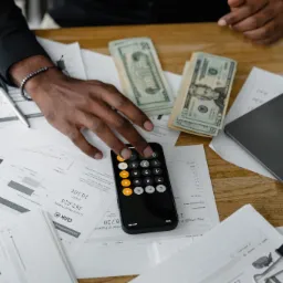 Osoba używa kalkulatora w telefonie nad stosem dokumentów z wykresami, obok laptop i pliki banknotów.