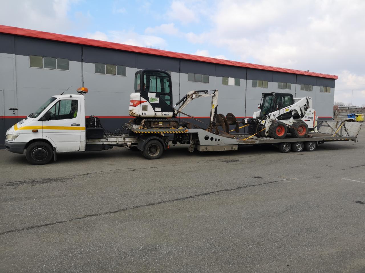 Dwuosiowa laweta z białym ciągnikiem Mercedes i pomarańczowym światłem ostrzegawczym, przewożąca minikoparkę Bobcat E16 i miniładowarkę Bobcat na tle szarego budynku z czerwoną linią dachu.