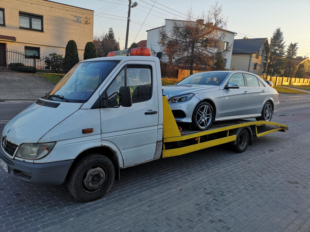 Biała laweta z żółtymi elementami transportuje srebrny samochód osobowy na tle zabudowy jednorodzinnej.