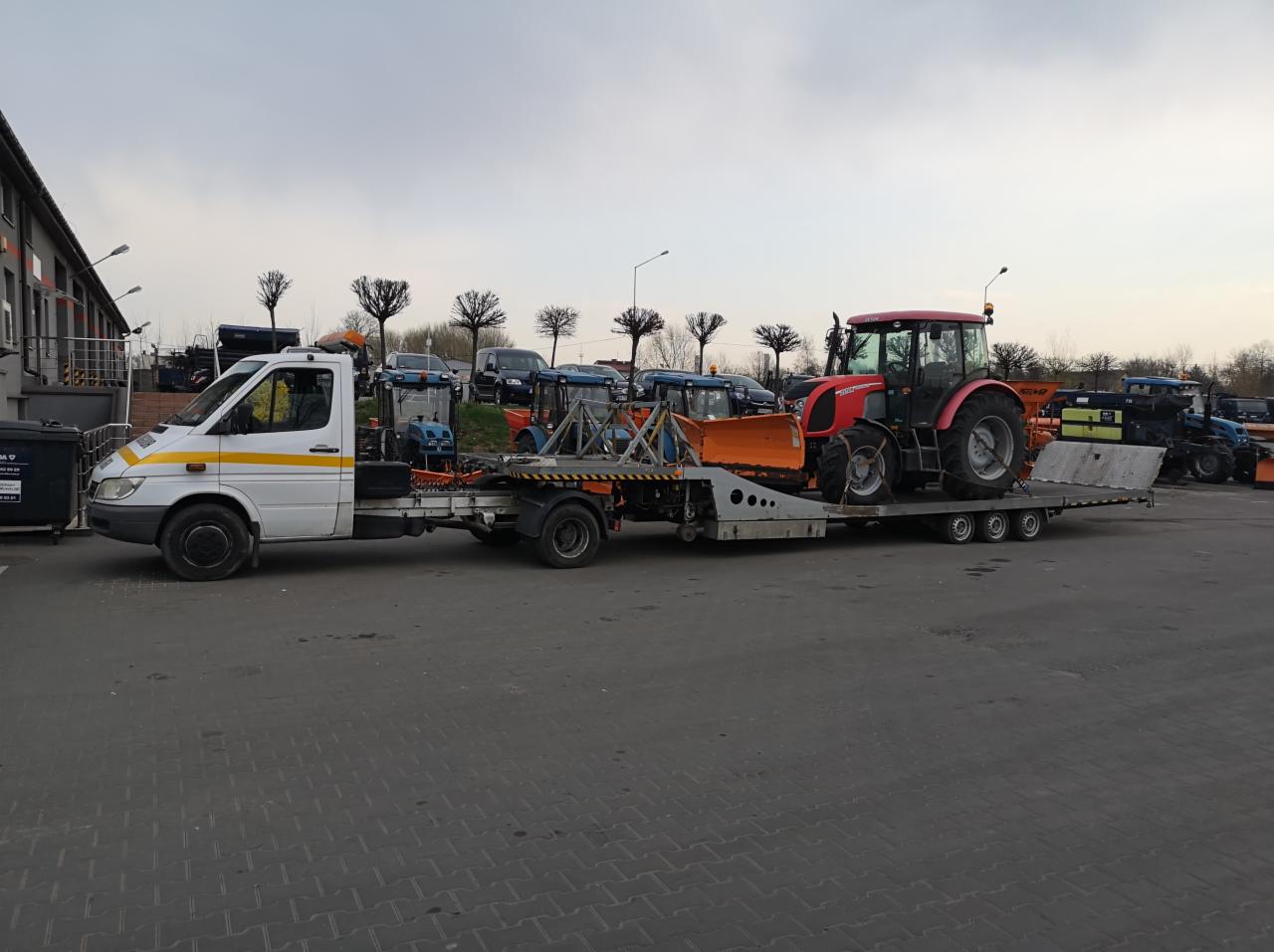 Biała laweta z żółtym pasem transportuje czerwony traktor z pomarańczowym pługiem na tle parkingu z zaparkowanymi traktorami.