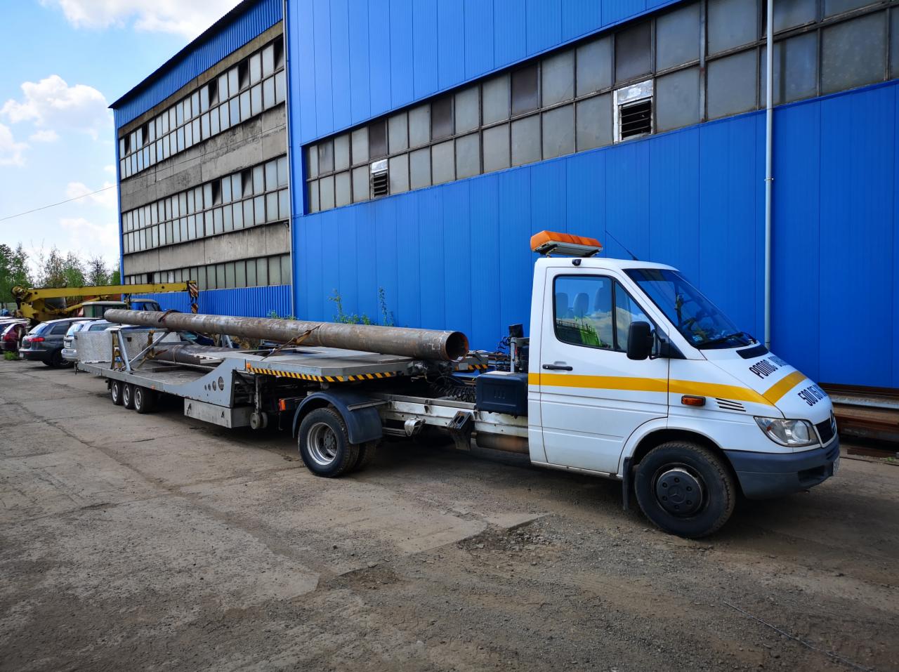 Biała laweta z żółtym pasem transportuje długą, szarą rurę na tle niebieskiego, przemysłowego budynku.