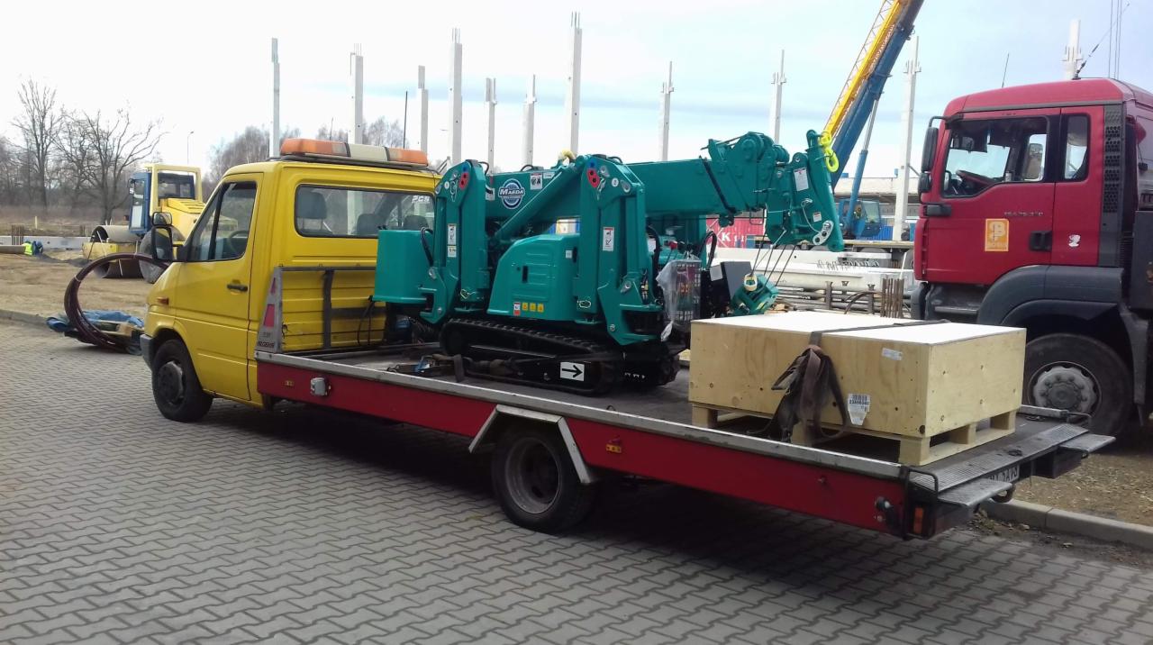 Żółta laweta z zielonym mini-dźwigiem gąsienicowym i drewnianą skrzynią na platformie, zaparkowana na kostce brukowej, w tle czerwona ciężarówka i plac budowy z żurawiem.