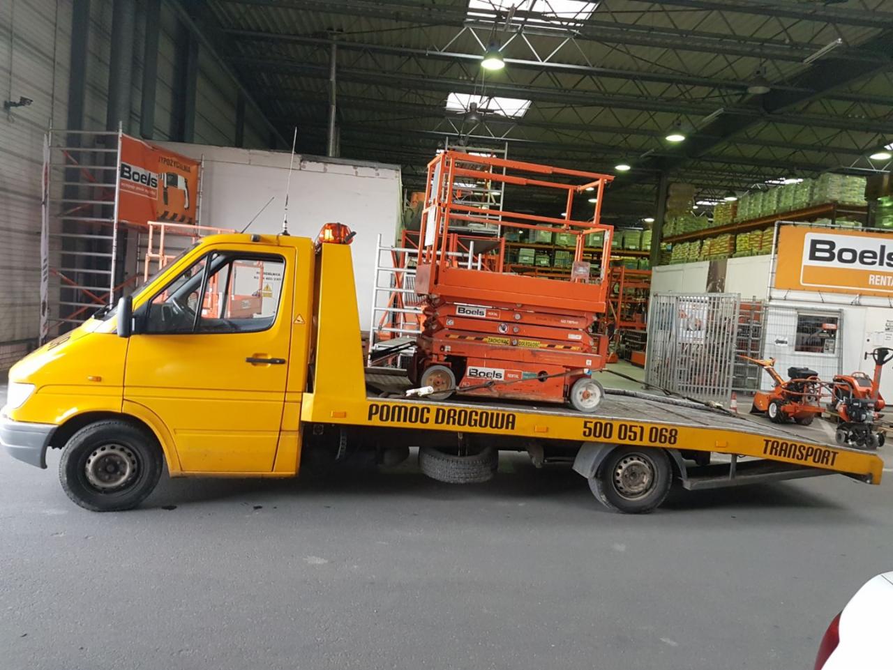 Żółta laweta z platformą roboczą marki Boels i drobnym sprzętem budowlanym na pace, zaparkowana w hali magazynowej.
