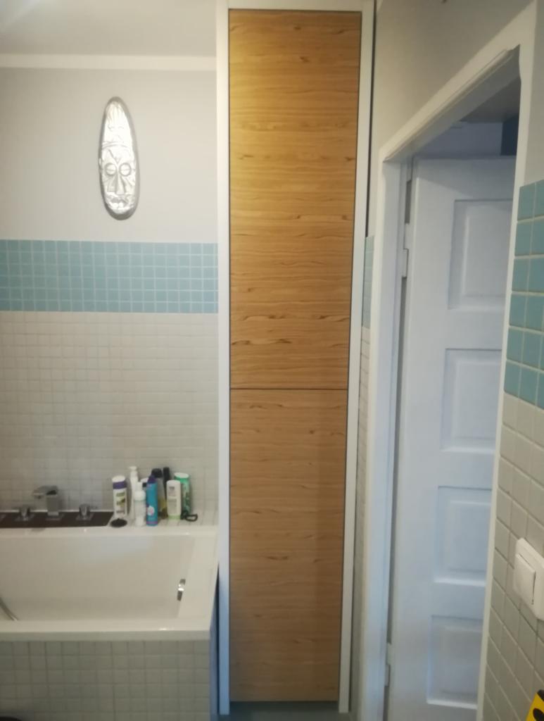 Wysoka szafa wnękowa z drewnianymi frontami w łazience z biało-błękitnymi płytkami i maską na ścianie.