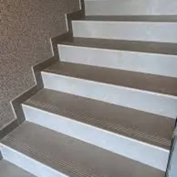 Schody wyłożone jasnymi, matowymi płytkami ceramicznymi z delikatnym wzorem antypoślizgowym na krawędziach stopni, widok z dołu, ściana z teksturą w tle.