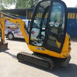 Żółta mini koparka JCB 8018 CTS z otwartą kabiną, stojąca na betonowym podłożu, z łyżką skierowaną w lewo, na tle samochodu dostawczego z reklamą i niebieskiego garażu.