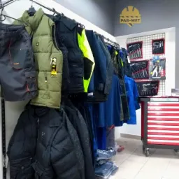 Ekspozycja odzieży roboczej w sklepie: szorty robocze z kieszeniami, kamizelka ocieplana khaki, kurtki puchowe i robocze, narzędzia GTools na ścianie i w wózku narzędziowym z czerwonymi szufladami,...