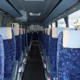 Wnętrze autobusu z rzędami niebieskich foteli z wzorem, widok z przodu na okno z budynkami w tle.