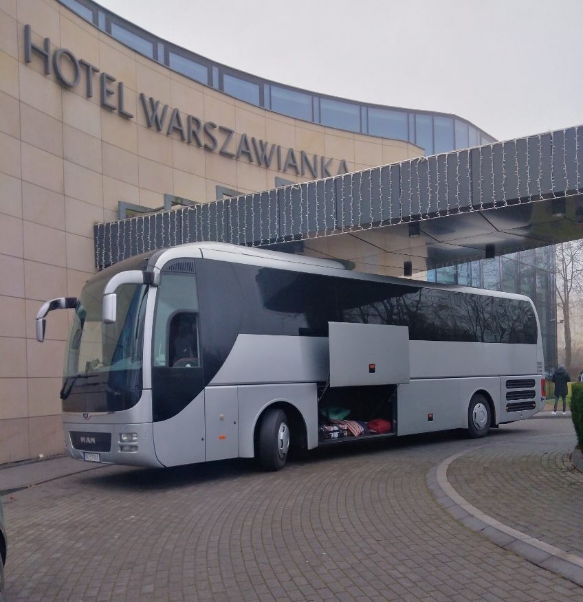 Srebrny autokar MAN z otwartym bagażnikiem przed hotelem Warszawianka. Widoczny fragment chodnika z kostki i sylwetka osoby w tle. Transport autokarowy.
