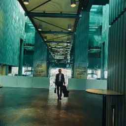 Mężczyzna w garniturze idzie przez przestronne, nowoczesne wnętrze z wysokimi kolumnami i przeszklonymi ścianami, trzymając płaszcz i torbę.