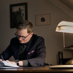 Kancelaria Adwokacka adw. Szymon Janiga - Adwokat w okularach, w eleganckiej marynarce, pisze przy biurku pod lampą w gabinecie. Na biurku leży otwarty notatnik i zegarek.