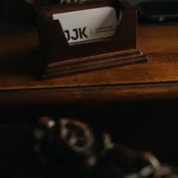 Wizytówka kancelarii adwokackiej JJ K w drewnianym podajniku na biurku, w tle książki i telefon. Ujęcie z bliska, ciemne, klimatyczne.