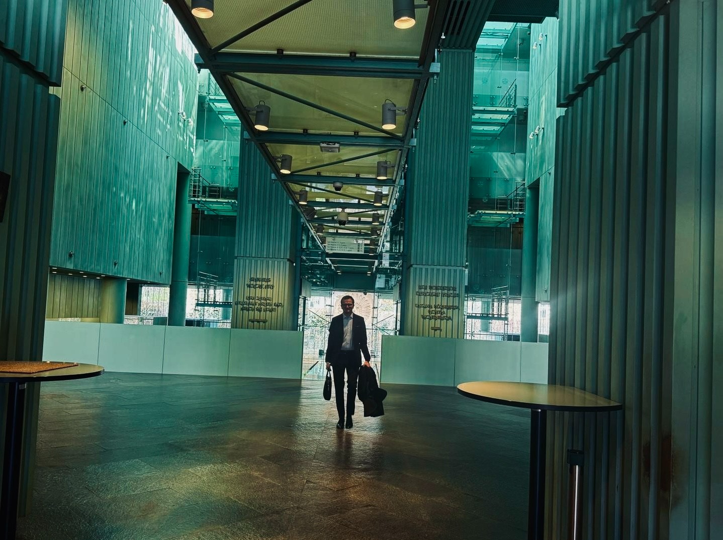 Mężczyzna w garniturze idzie przez przestronne, nowoczesne wnętrze z wysokimi kolumnami i przeszklonymi ścianami, trzymając płaszcz i torbę.