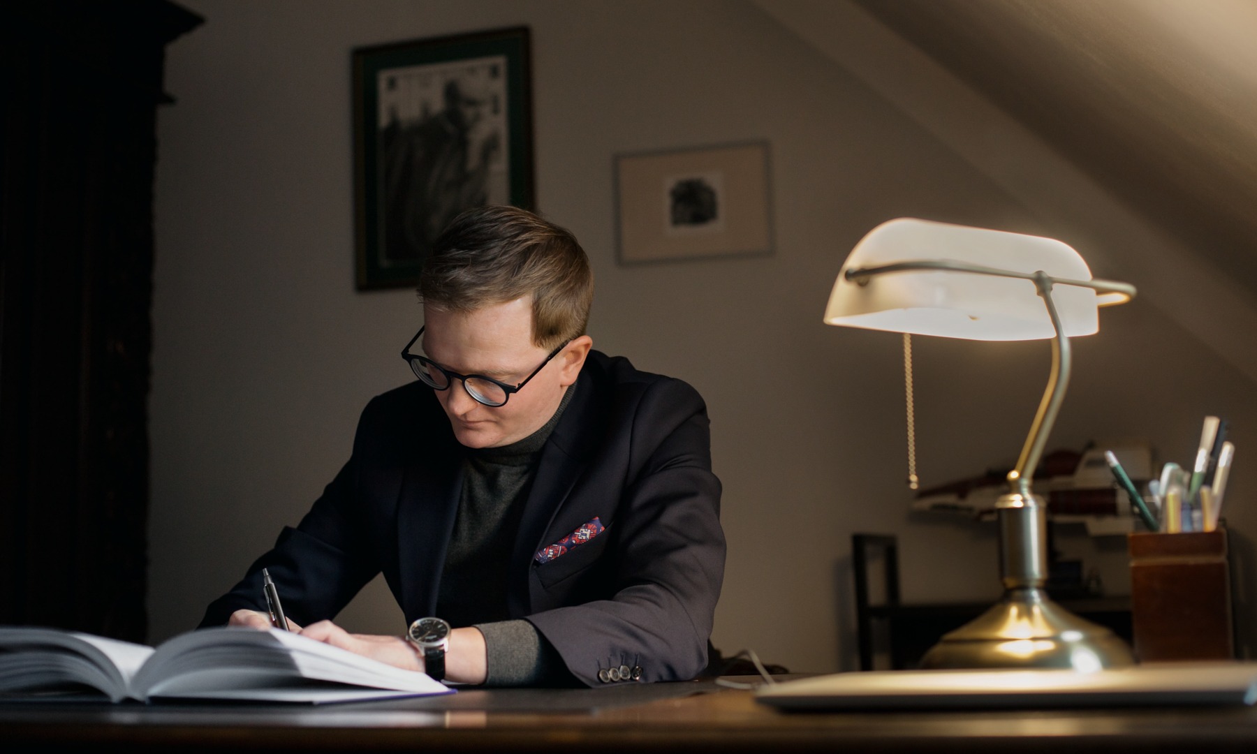 Adwokat w okularach, w eleganckiej marynarce, pisze przy biurku pod lampą w gabinecie. Na biurku leży otwarty notatnik i zegarek.