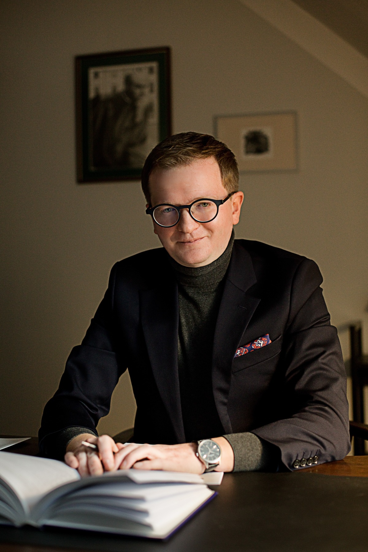 Portret mężczyzny w okularach siedzącego za biurkiem z otwartą książką i długopisem, w marynarce z ozdobną poszetką. Stonowana kolorystyka, ciepłe oświetlenie.