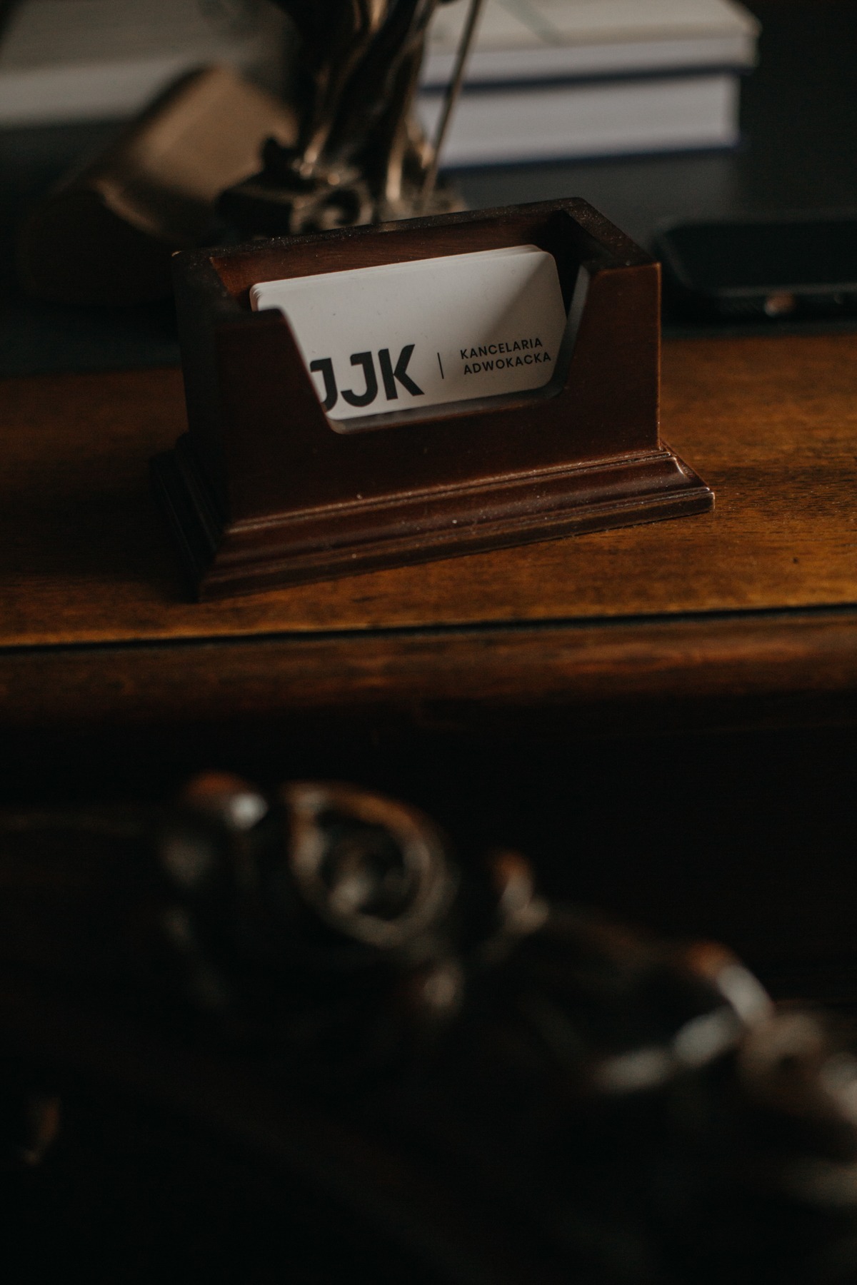 Wizytówka kancelarii adwokackiej JJ K w drewnianym podajniku na biurku, w tle książki i telefon. Ujęcie z bliska, ciemne, klimatyczne.