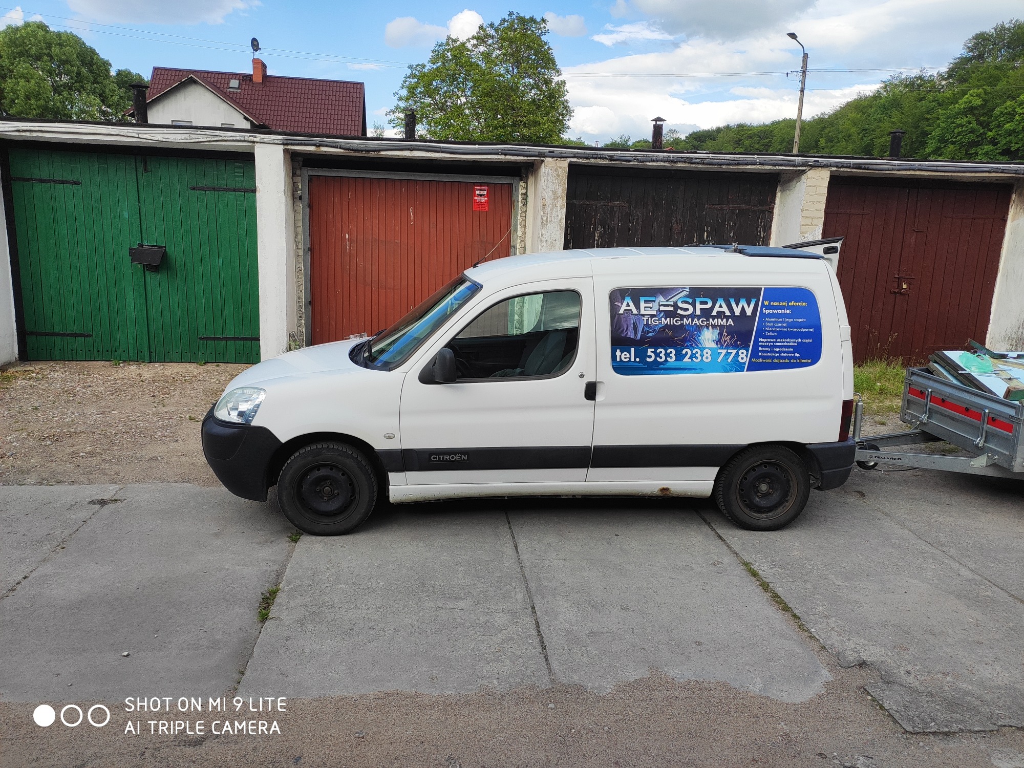 Biały samochód dostawczy z reklamą firmy spawalniczej AE-SPAW, zaparkowany przed garażami z zielonymi i brązowymi drzwiami.