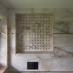 Wnęka w łazience wyłożona marmurowymi płytkami z dekoracyjnym wzorem w kratkę, wykończona złotymi listwami, obok okno z ciemną ramą.