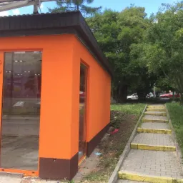 Pomarańczowy budynek z wykończoną elewacją, brązowym cokołem i przeszklonymi drzwiami, widoczne schody prowadzące do budynku i zieleń w tle.