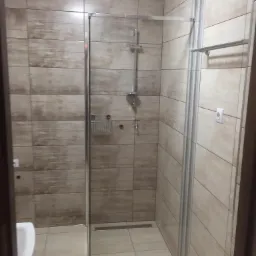 Wykończona kabina prysznicowa z beżowymi płytkami imitującymi drewno, szklane drzwi prysznicowe z chromowanymi detalami, widoczny odpływ liniowy w podłodze i biały włącznik światła na ścianie.