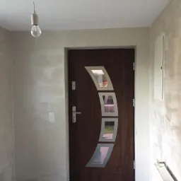 Wnętrze po remoncie: ciemne drzwi z trzema podłużnymi okienkami, ściany pokryte jasną, nieregularną gładzią, surowa żarówka zwisająca z sufitu.