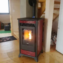 Ciemnoczerwony piec wolnostojący na pellet z dekoracyjnymi ornamentami, płomień widoczny przez szybę, ustawiony w drewnianym wnętrzu z widocznymi schodami i oknem.