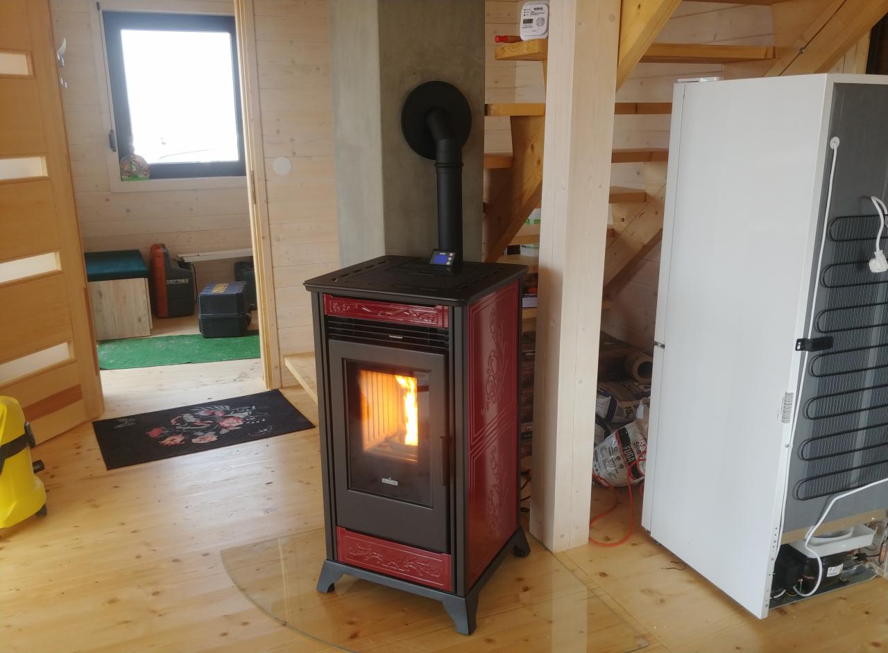 Ciemnoczerwony piec na pellet z płomieniami w palenisku, zainstalowany w drewnianym domu z widocznymi schodami i lodówką.