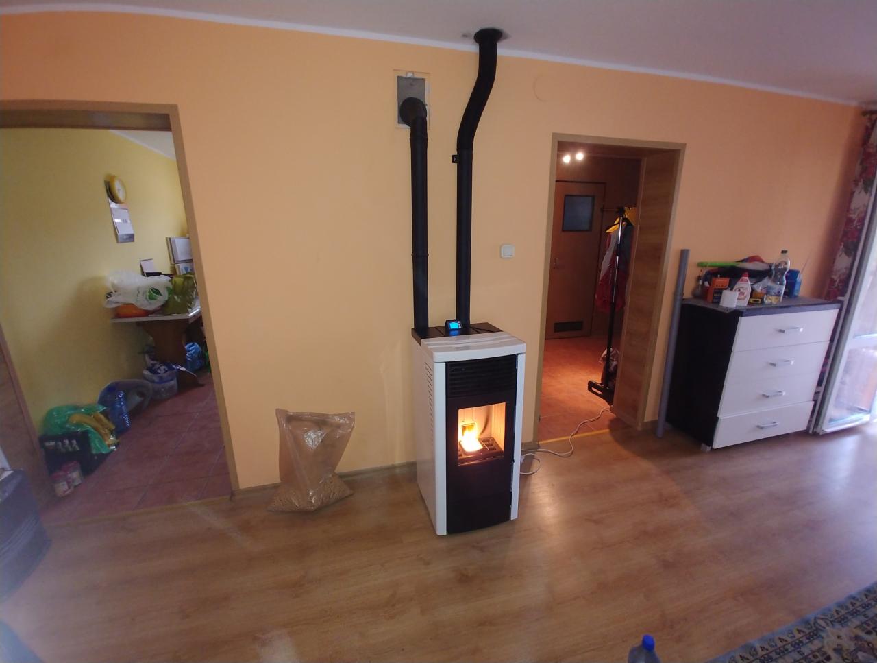 Nowoczesny piec na pellet z czarnym kominem w jasnym pokoju, obok worek z pelletem, widoczne płomienie w palenisku.