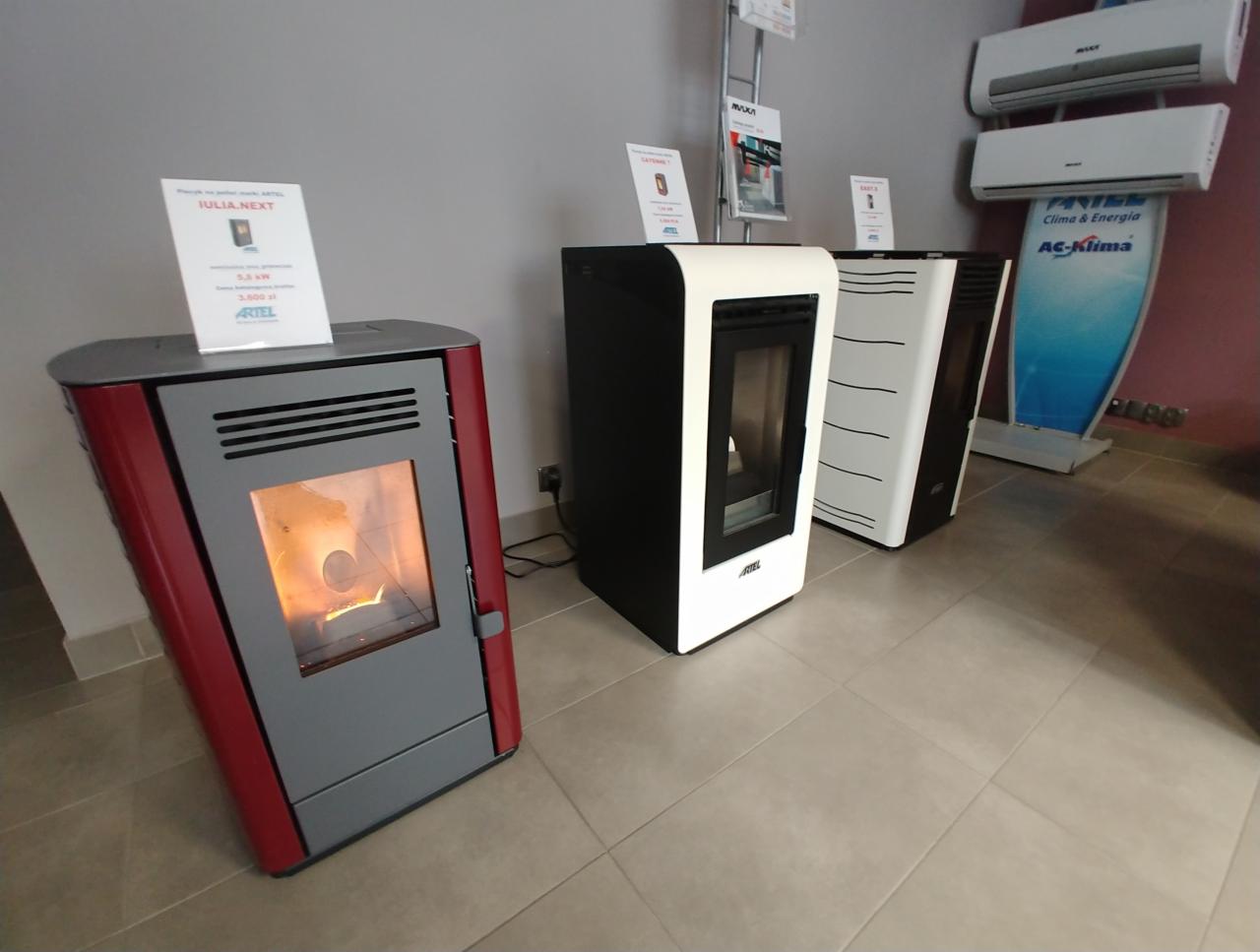 Wystawa nowoczesnych pieców na pellet w salonie, model w kolorze czerwono-szarym z płomieniem wewnątrz, obok modele czarno-białe i klimatyzatory na ścianie.