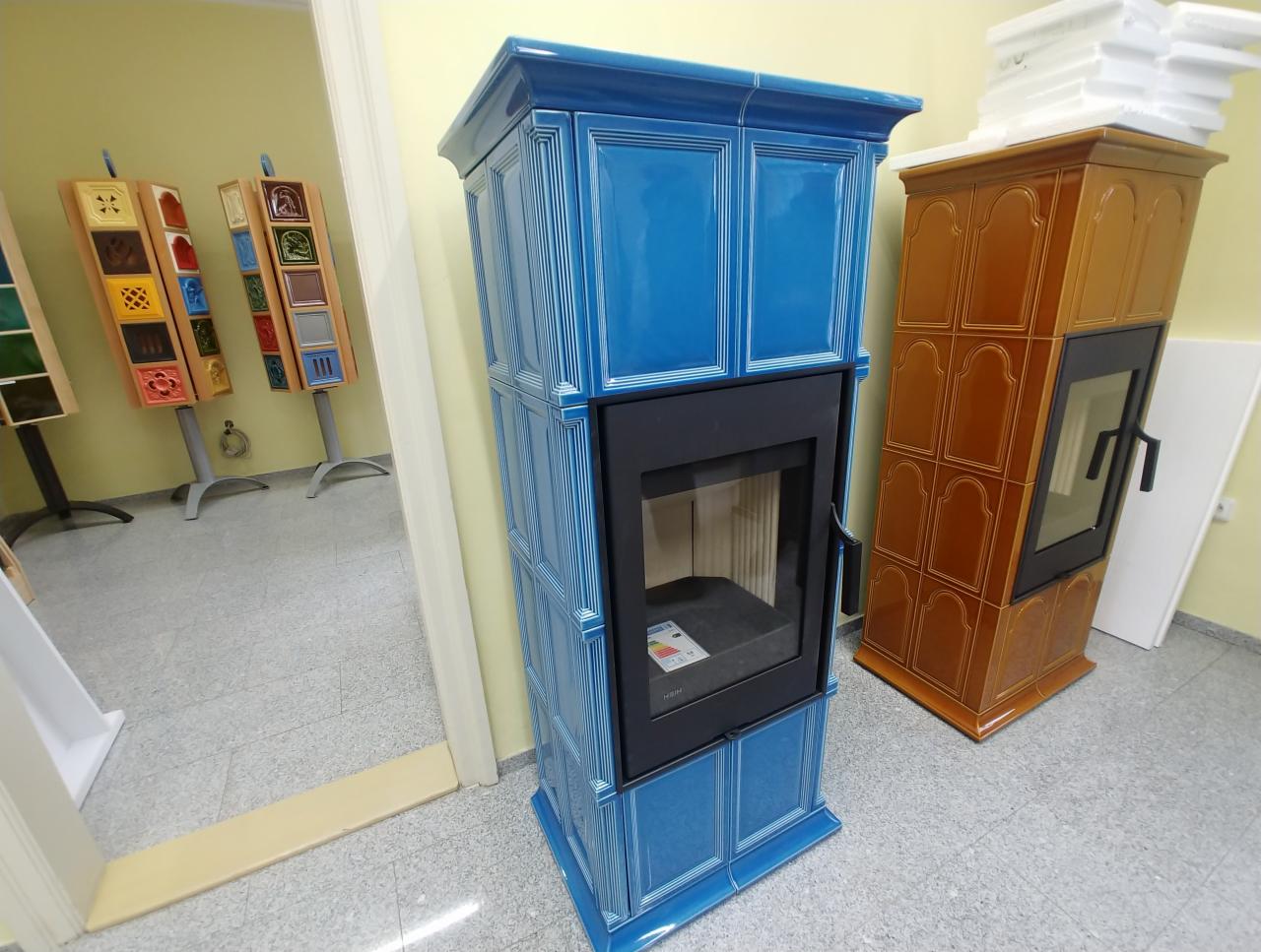Niebieski, kaflowy kominek wolnostojący z czarnym wkładem, prezentowany obok brązowego pieca kaflowego w jasnym pomieszczeniu. W tle ekspozycja płytek ceramicznych na stojakach.