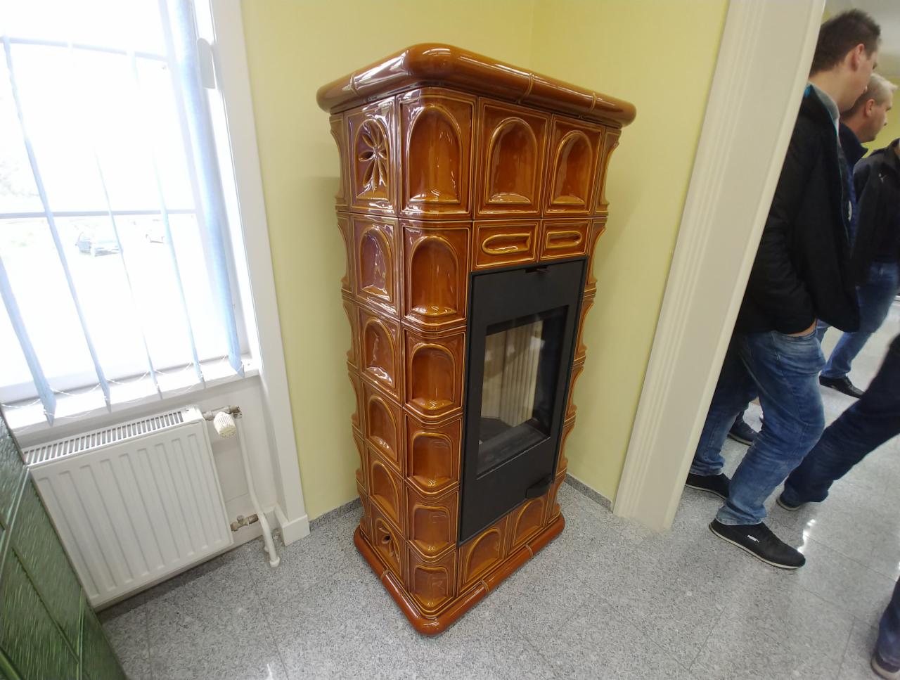 Glazowany piec kaflowy w kolorze miodowym z czarnym wkładem kominkowym, ustawiony w rogu pokoju z oknem i grzejnikiem, widoczni ludzie w tle.