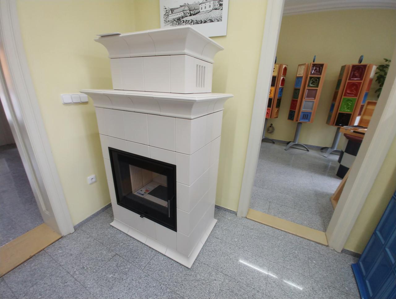 Biały, kaflowy piec z czarnym wkładem kominkowym, umieszczony w rogu pokoju z jasnymi ścianami i szarą podłogą. W tle, przez otwarte drzwi, widoczne ekspozytory z kolorowymi kaflami.