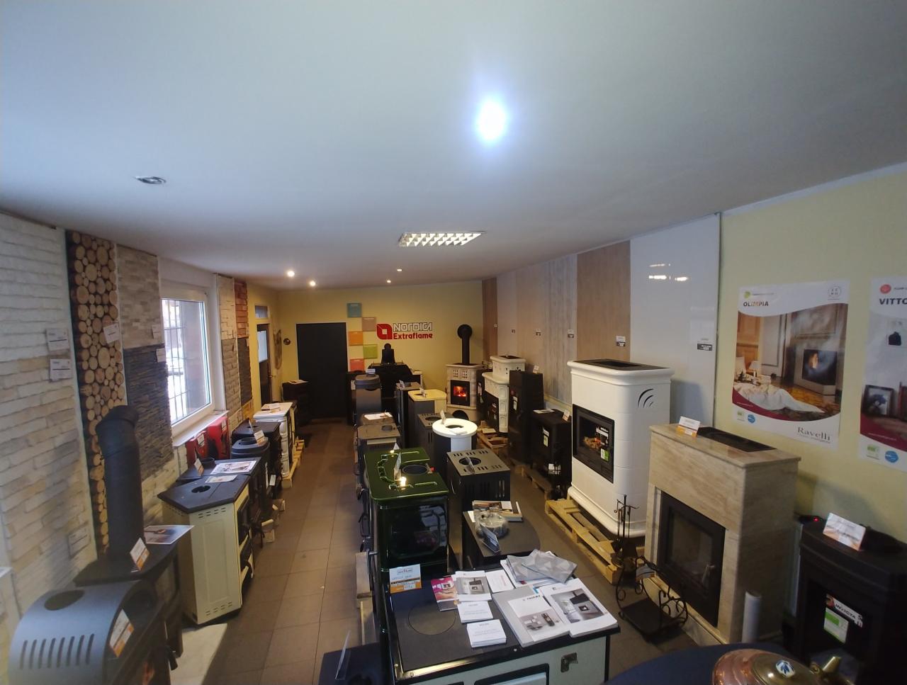 Ekspozycja różnorodnych pieców i urządzeń grzewczych w salonie, widoczne modele wolnostojące i z obudową kamienną, na ścianach plakaty reklamowe producentów.