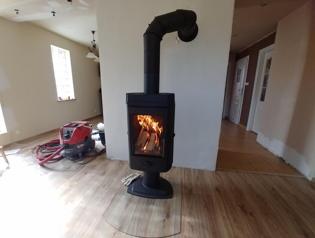 Czarny, wolnostojący kominek z palącym się drewnem, zamontowany na szklanej płycie ochronnej, widoczny odkurzacz przemysłowy w tle, podłoga laminowana imitująca drewno.