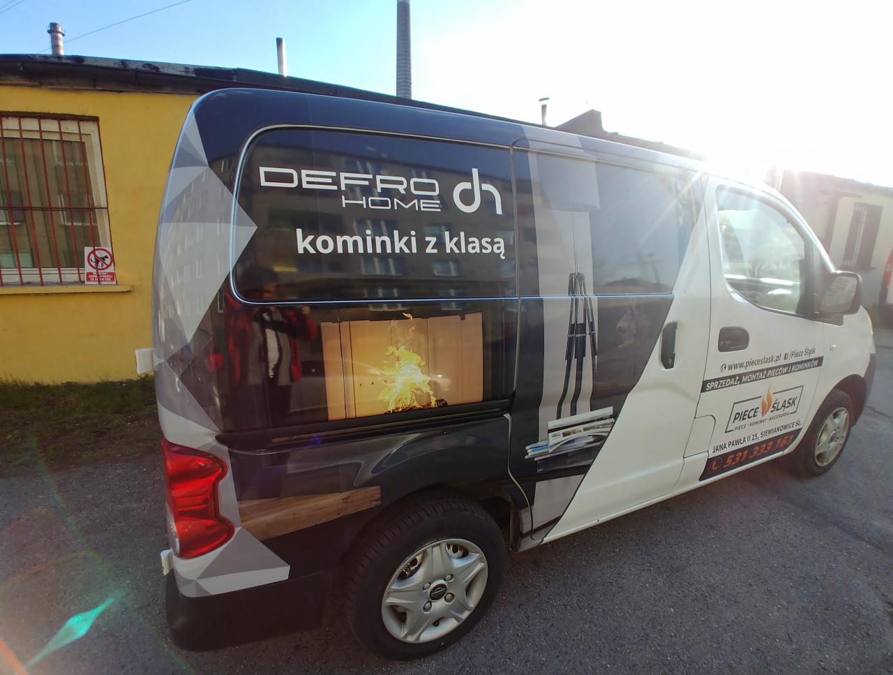 Okolicznościowy samochód dostawczy z reklamą firmy Defro Home, prezentujący wizualizacje wkładu opalanego drewnem oraz nowoczesnego pieca.