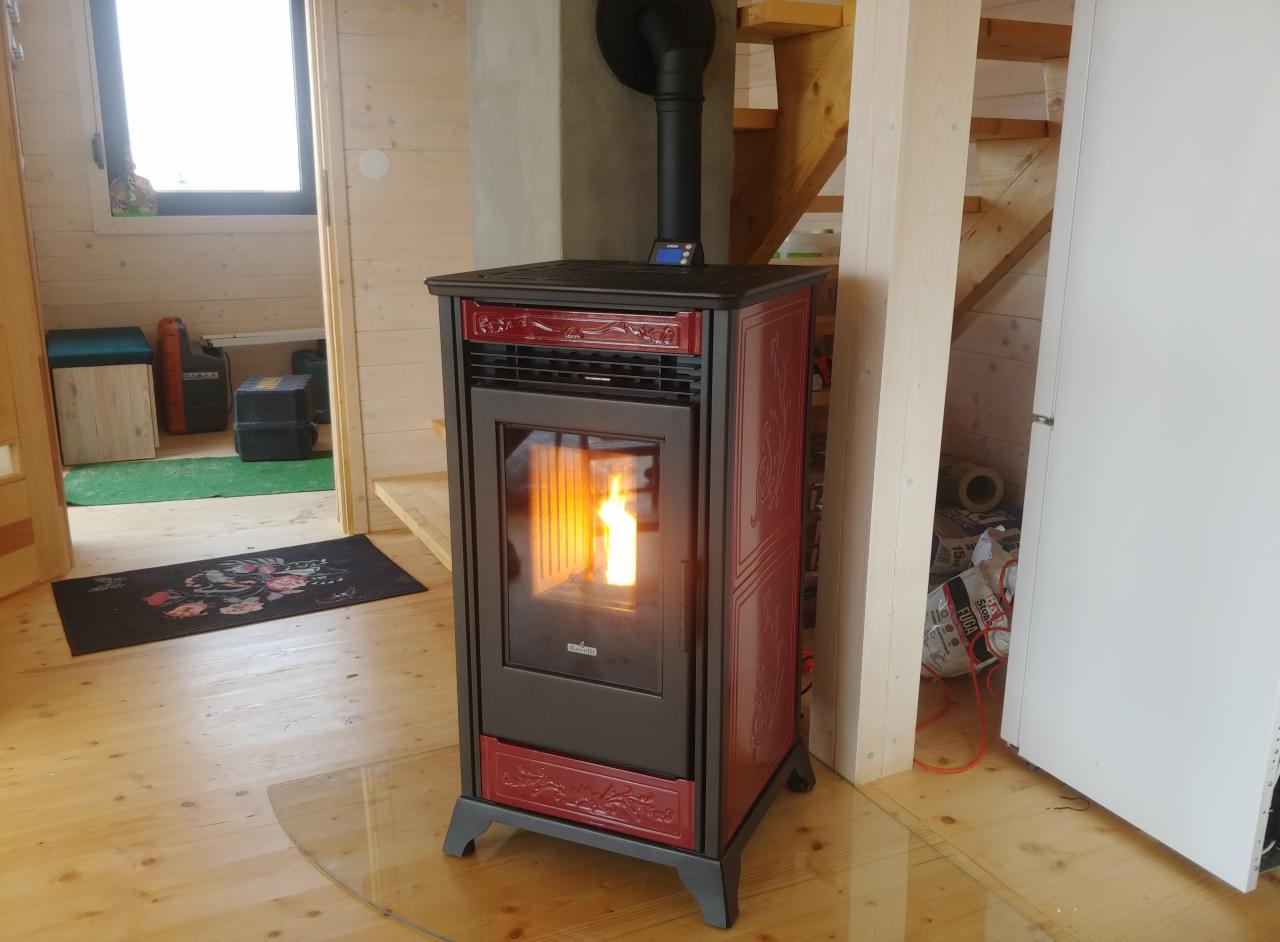 Ciemnoczerwony piec wolnostojący na pellet z dekoracyjnymi ornamentami, płomień widoczny przez szybę, ustawiony w drewnianym wnętrzu z widocznymi schodami i oknem.