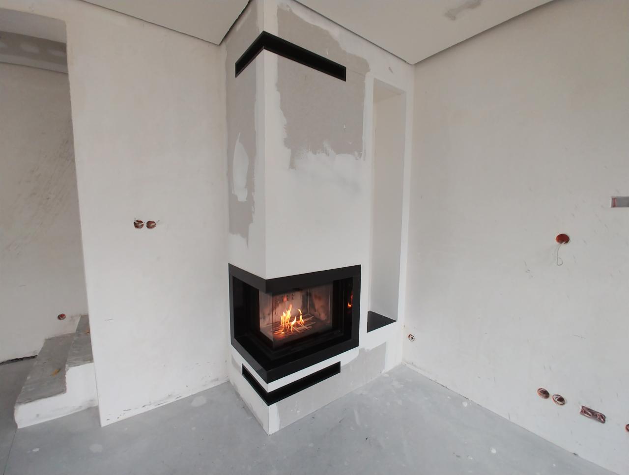 Narożny wkład z paleniskiem w trakcie montażu, obudowa w surowym stanie, widoczne ślady po szpachlowaniu na ścianach i podłodze.
