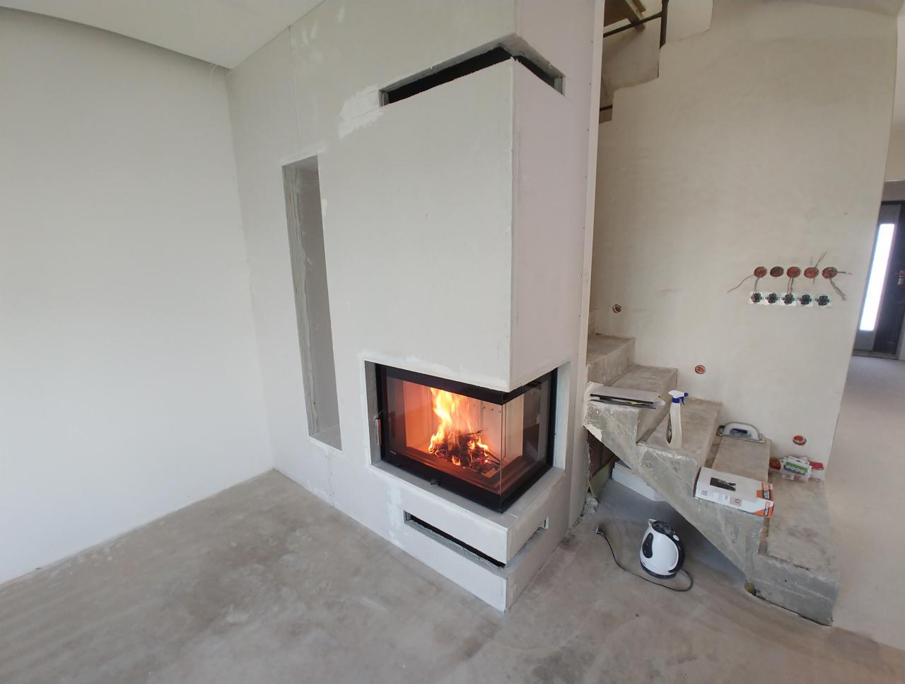Narożny kominek z paleniskiem w surowym wykończeniu, widoczne elementy instalacji elektrycznej i schody w trakcie budowy.