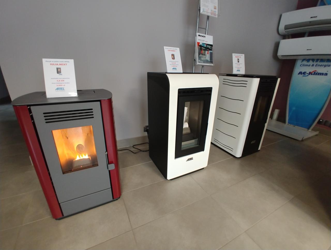 Wystawa trzech różnych modeli pieców na pellet marki Artel: czerwono-szary z płomieniem wewnątrz, czarno-biały i biało-czarny z poziomymi paskami, na tle płytek podłogowych i klimatyzatorów.