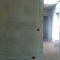 Surowa ściana z betonu komórkowego z poprowadzonymi natynkowo przewodami elektrycznymi w białej rurce PCV i czerwonymi puszkami gniazdek w budynku w stanie deweloperskim.