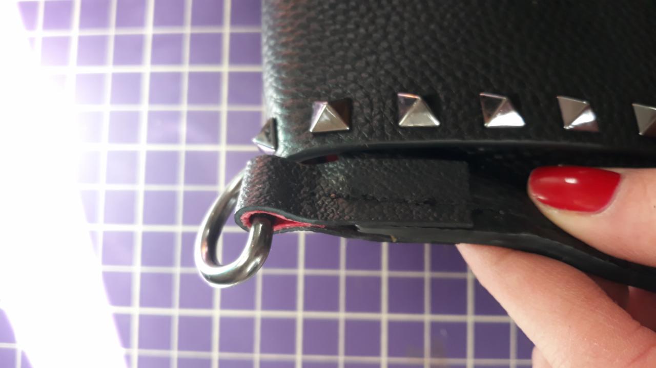 Fragment czarnej skórzanej torebki z metalowymi ćwiekami i uchwytem w kształcie pierścienia, trzymanej w dłoni z czerwonym paznokciem, na tle fioletowej kratownicy.