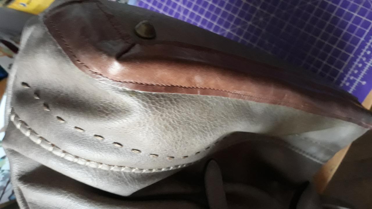 Szaro-brązowa skórzana torba z ozdobnymi przeszyciami i ciemnobrązowym wykończeniem, widoczna na tle fioletowej maty do cięcia.