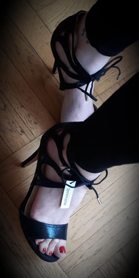 Dwie stopy w czarnych, eleganckich sandałach na wysokim obcasie z wiązaniami, na parkiecie; na jednym bucie widoczna metka marki Mariamare.