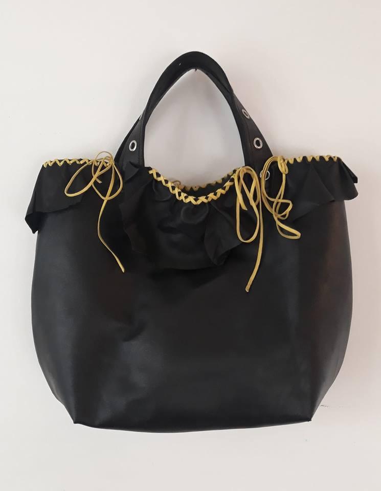 Czarna torebka shopper z falbaną i złotymi sznurkami, wisząca na białej ścianie.