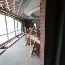 Wnętrze w trakcie prac wykończeniowych: surowa ściana z cegły, okna z dużymi przeszkleniami, konstrukcja sufitu podwieszanego i drewniane rusztowanie.