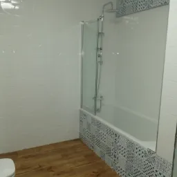 Wykończona łazienka we Wrocławiu z drewnopodobną podłogą, białą armaturą i płytkami w geometryczne wzory nad wanną z parawanem.